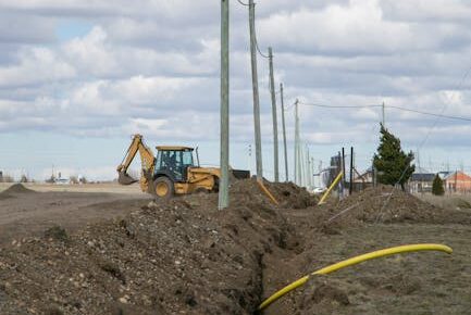 A backhoe digging at a construction site with utility poles and overhead wires under a cloudy sky.